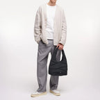 A male model holding a black Lieblingstasche wavy puffer shoulder bag while wearing a beige cardigan and grey trousers.