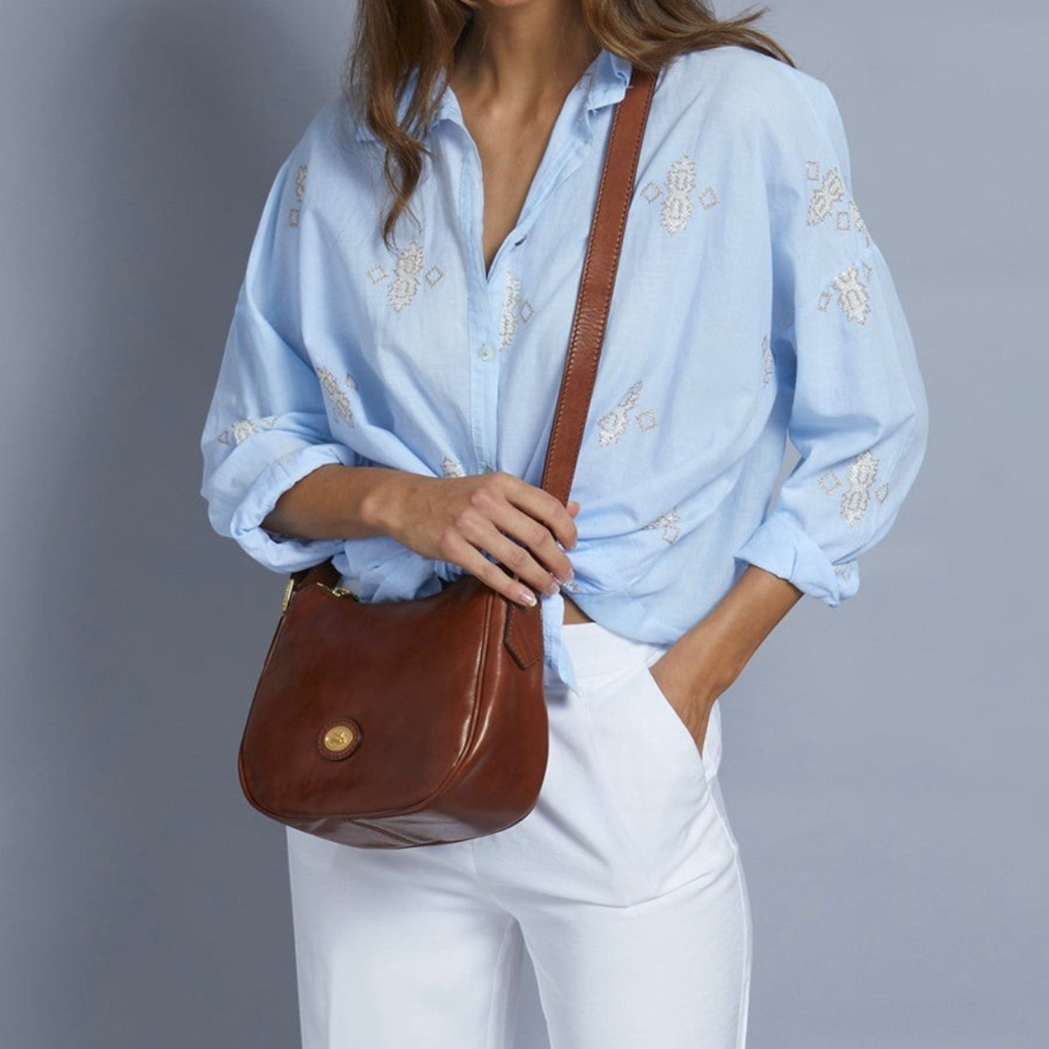 Lieblingstasche brown leather crossbody bag with a gold buckle and zipper closure, worn by a model in a blue embroidered shirt and white pants.
