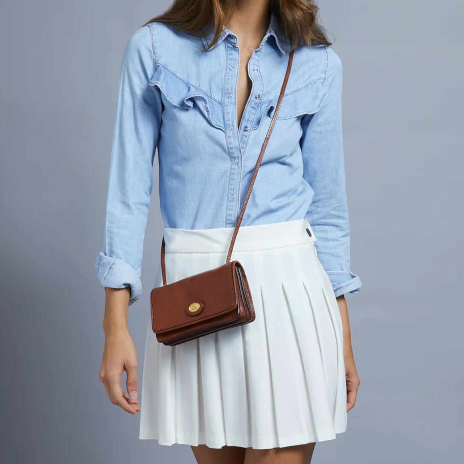 Lieblingstasche brown leather mini crossbody bag with gold clasp, worn by a model in a denim shirt and white skirt.