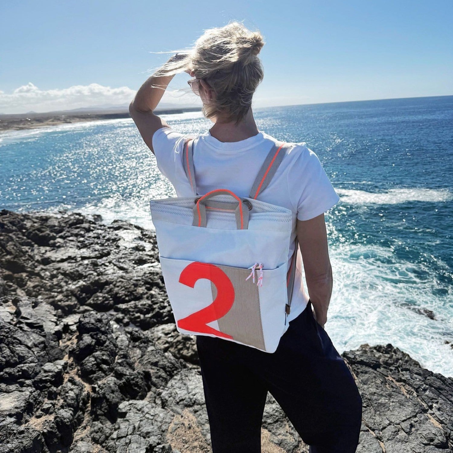 Lieblingstasche Kaje white canvas backpack with tan accents and a large orange number 2 graphic, worn by a woman overlooking the ocean.