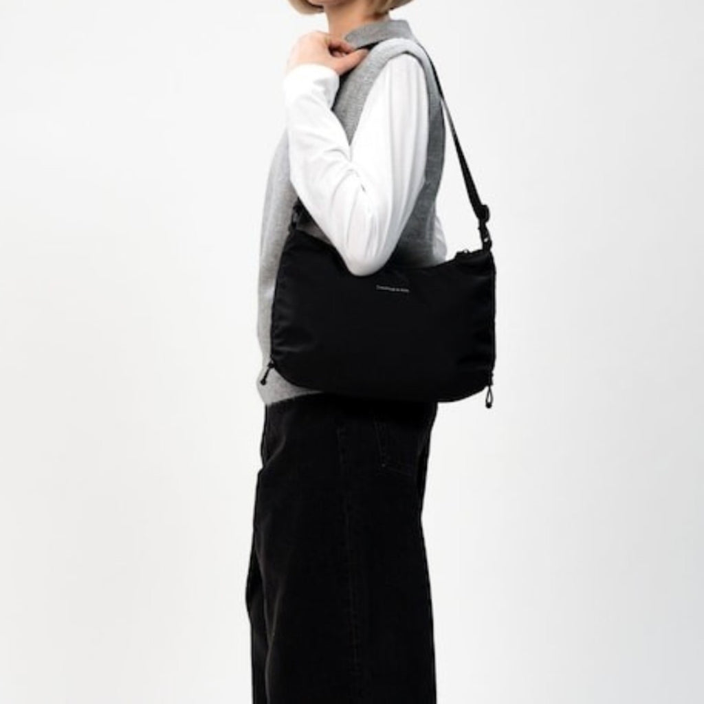 Lieblingstasche Kapten & Son Skara Small black shoulder bag worn by a woman in a grey vest and white long-sleeve top.