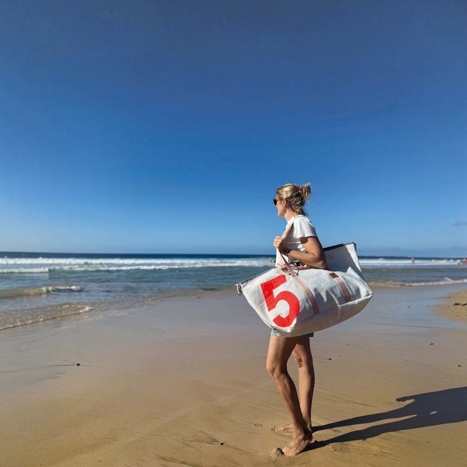 Woman carrying a white Lieblingstasche Kutter XL sailcloth beach bag with a large red number 5 on a sunny beach