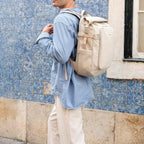 Man wearing a beige Lieblingstasche Lisbon Pro travel backpack with roll-top design against a blue tiled wall