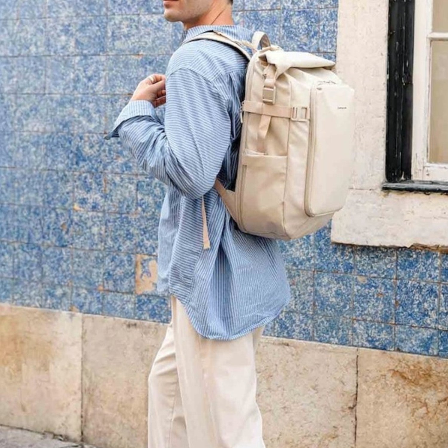 Man wearing a beige Lieblingstasche Lisbon Pro travel backpack with roll-top design against a blue tiled wall