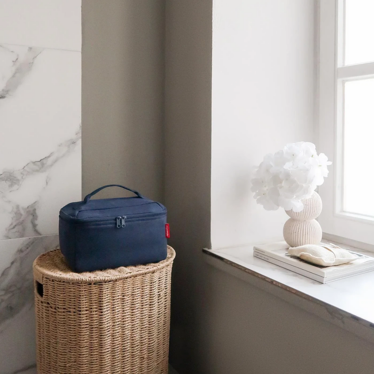 Lieblingstasche navy blue beautycase sitting on a woven wicker basket in a bright, minimalist bathroom setting